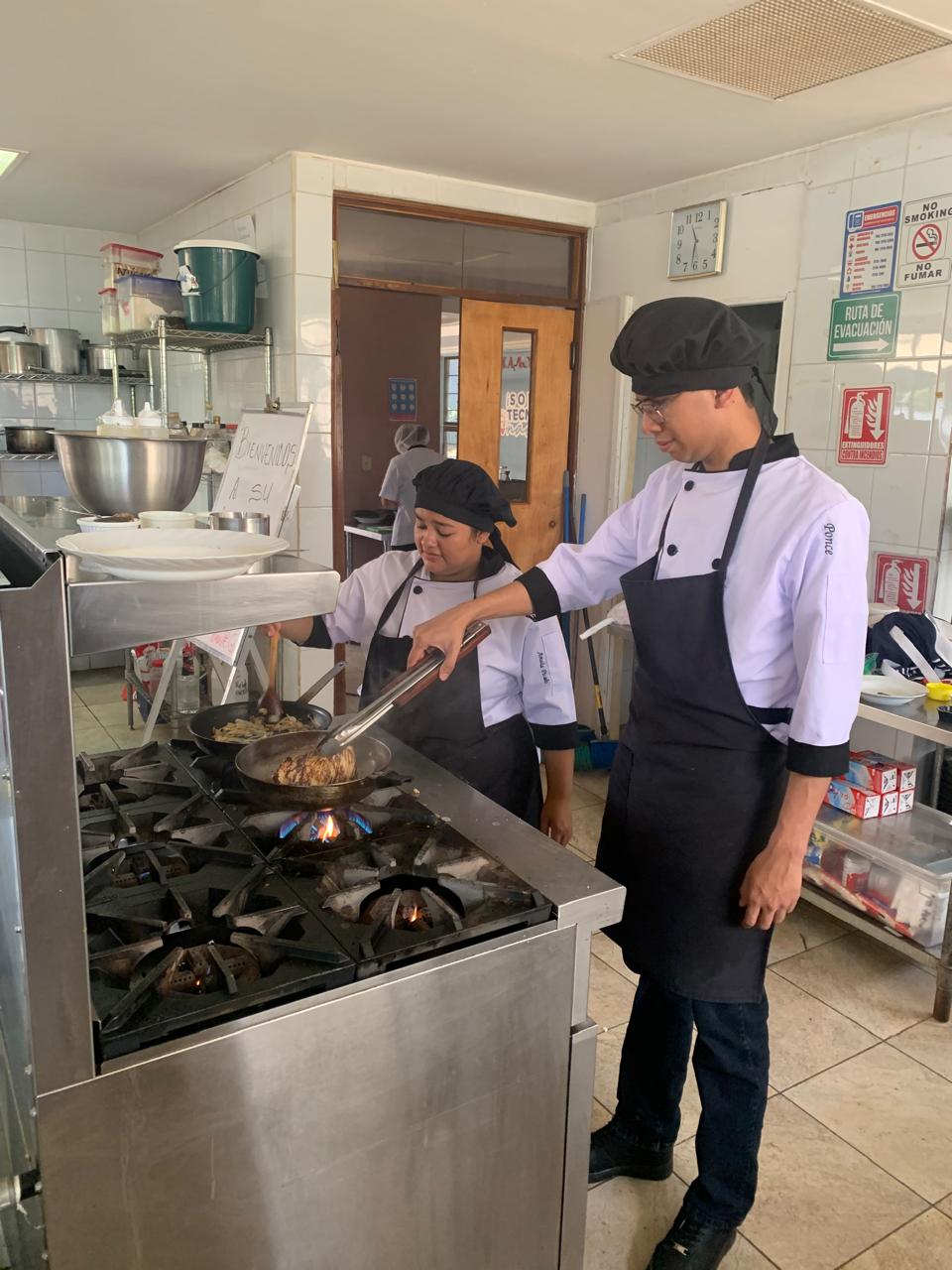 Dos estudiantes de cocina con uniforme preparan un platillo en una cocina didáctica con estufa de gas